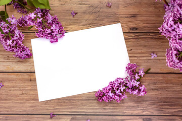 White blank sheet of paper with blooming lilac flowers on wooden background. Invitation or greeting card for Valentine's Day and Mother's Day. Top view, flat lay, mock up, copy space. Spring concept.