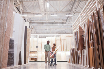 Wide angle portrait of African-American father and son shopping together in hardware store standing with cart in wood and boards isle, copy space