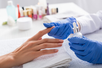Obraz premium partial view of manicurist holding vial of cuticle remover while touching finger of client, blurred background