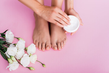 Obraz na płótnie Canvas cropped view of barefoot woman holding cosmetic cream near white eustoma flowers on pink background