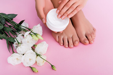 partial view of barefoot woman holding cosmetic cream near white eustoma flowers on pink background