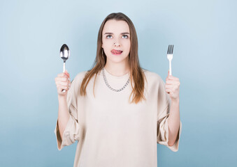 A hungry girl holds a fork and a spoon in her hands, licks her lips and thinks about delicious food on a blue background. Diet and food concept.