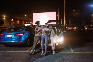 Full length shot of three cheerful young friends looking at camera while standing together by the car parked in front of a big white screen to watch movies