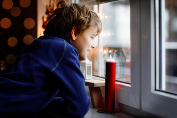 Happy adorable kid boy sitting near window and looking outside on snow on Christmas day or morning. Smiling healthy child fascinated observing snowfall and big snowflakes