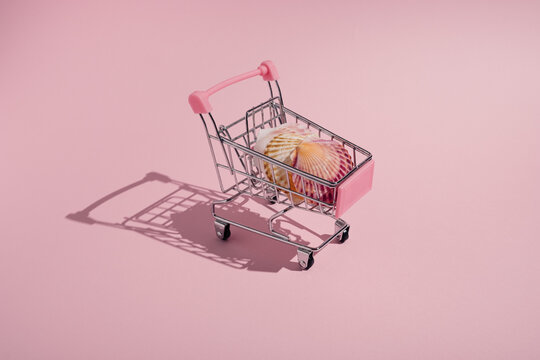 Creative summer concept made of shopping cart and colorful sea shells on pastel pink background.