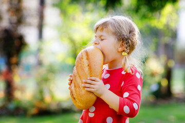 Little toddler girl holding big loaf of bread. Funny happy child biting and eating healthy bread,...