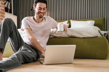 Laughing man using laptop and drinking coffee while sitting on floor
