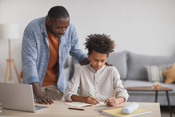 Portrait of proud African-American father helping teenage boy doing homework at desk in modern home...