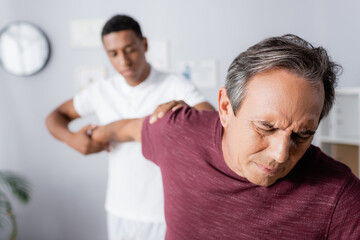 mature man suffering from pain while receiving massage from african american physiotherapist on blurred background