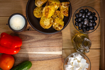 Rustic lunch with baked potatoes and yogurt in ceramic dishes on a wooden table. In addition, there are black olives, olive oil, feta cheese, tomato, sweet pepper, cucumber on the table.