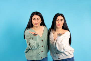 Portrait of twin girls who are sending an air kiss on a blue background in the same clothes.