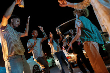 A close up of a good-looking group of young friends of different nationalities dancing in front of each other outside on a parking site while eating pizza and drinking beer