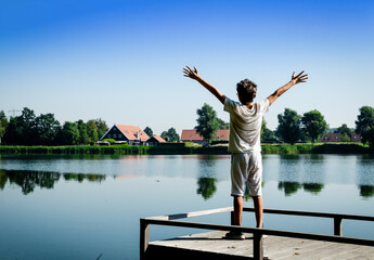 Fototapeta na wymiar Netherlands, Goes. Dutch countryside lake landscape: reeds alternating with wooden piers. A Caucasian boy is inspired by nature and raises his arms to the sky.
