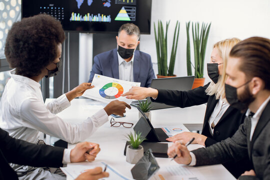 Multiracial high-skilled diverse businesspeople in black face masks, sitting around boardroom table and discussing business strategy. African and Caucasian women holding paper with infographics