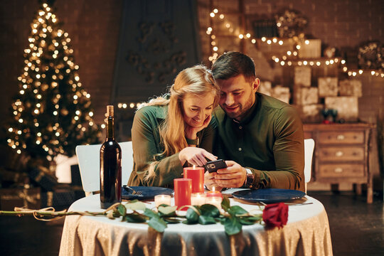 Sits and using phone. Young lovely couple have romantic dinner indoors together