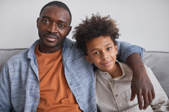 Horizontal Waist Up Portrait Shot Of Happy African American Man Sitting On Sofa With His Teen Son Looking At Camera