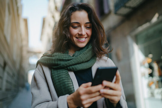 Portrait of young smiling dark-haired woman wearing light-colored coat and green scarf, walking city streets and using smartphone to interact with her friends. Good vibes. Modern technologies concept