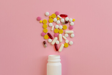Assorted medical pills and capsules scattered from a white plastic bottle on a pink background.