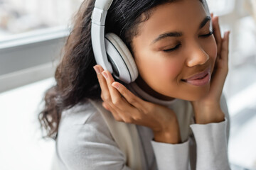 dreamy african american woman listening music in wireless headphones with closed eyes, blurred background