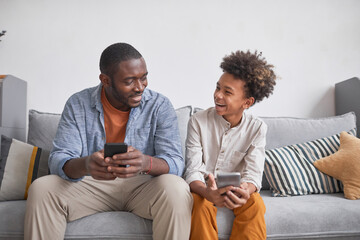 Joyful teenager having fun with his father playing video game on smartphones together while staying...