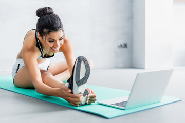 cheerful african american woman stretching while sitting on fitness mat near laptop