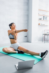 Fototapeta na wymiar happy african american woman stretching on fitness mat near laptop at home