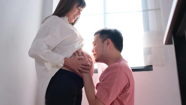 Asian man or husband kiss belly of his pregnant wife in front of glass windows with morning light and they express love and care together with bonding and cheerful in family concept.
