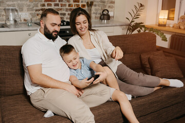 A dad, a son, and a mom on the sofa. A happy child is playing in the game on the smartphone between a father and a mother in the evening at cozy home.