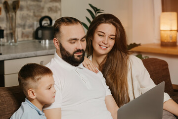 A close up photo of father with a beard who is showing his achievements at work on a laptop to his son and smiling wife at home. Dad is working remotely on a computer between relatives in the evening