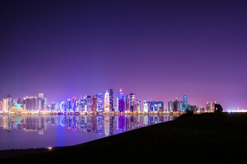 Fototapeta na wymiar (Selective focus) Stunning panoramic view of the Doha skyline illuminated at dusk in the distance and some people relaxing on a hill in the foreground.