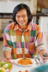 Happy Asian middle-aged man eating plate of delicious spaghetti and listening to friends at festive dinner