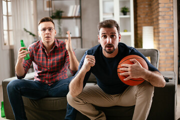 Two young friends enjoying at home. Men drinking beer and watching sports game on tv.