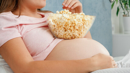 Closeup of pregnant woman lying in bed and eating popcorn from bowl lying on her big belly