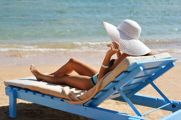 Beautiful woman sunbathing on a beach at tropical travel resort, enjoying summer holidays. Girl holding a hat with her hands on sun lounger near sea