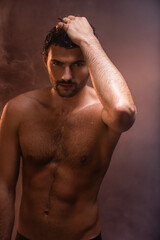 Fototapeta na wymiar sexy, wet man with muscular torso posing with hand on head on dark background with smoke