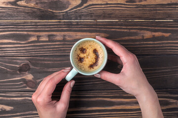 Female hands holding a cup of coffee with foam over a wooden table. with chocolate sprinkles