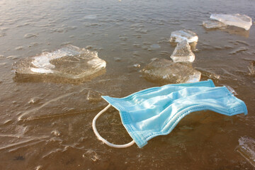 Fototapeta na wymiar Blue protective mask on frozen river. Discarded pandemic face mask on ice in winter. Top view