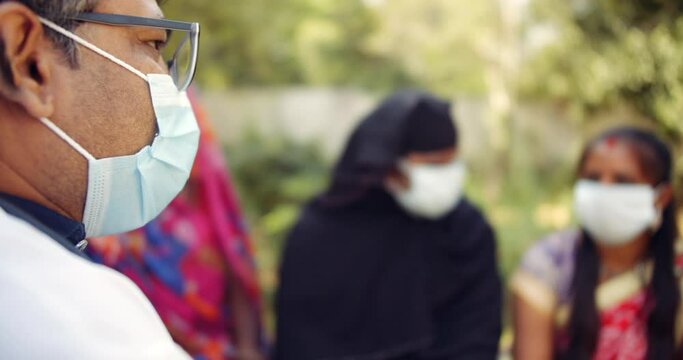 Shallow focus bokeh of close-up face of a doctor wearing face mask outdoors in nature public park informs a group of people wearing colorful vibrant traditional dress about coronavirus covid-19