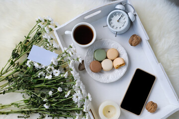 Fototapeta na wymiar white wooden tray with black tea, coffee, plate with macaroon cakes, alarm clock, flowers, blank screen phone, concept of technology, time, joys of life, flatley, top view