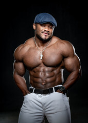 Fototapeta na wymiar Sexy afro american bodybuilder with no shirt. Athlete poses to the camera in white jeans and blue cap. Strong muscular body.