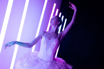 Fototapeta premium Portrait of a ballerina in a white tutu making a beautiful pose. A graceful ballet dancer gracefully poses against the backdrop of bright neon lights in a dark studio.