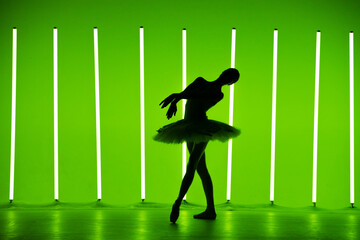 Beautiful young ballerina dancing classical ballet in a dark studio against a background of bright green neon lights. Silhouette of a slender female dancer in a white tutu and pointe shoes. © kinomaster