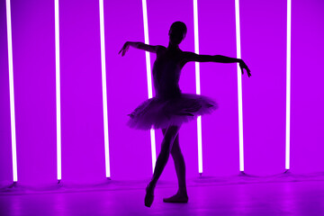 Fototapeta na wymiar Young classical ballet dancer posing in a dark studio against a background of purple neon lights. Slender ballerina is silhouetted on tiptoes in white tutu. Ballet school poster.