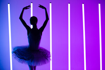 Young graceful ballerina in pointe shoes and a white tutu demonstrates her dancing skills. A beautiful classical ballet dancer against the backdrop of bright neon lights in a dark studio. Silhouette. © kinomaster
