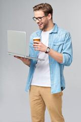 Concentrated young bearded man wearing glasses dressed in jeans shirt holding laptop and cup of coffee to go isolated over grey studio background