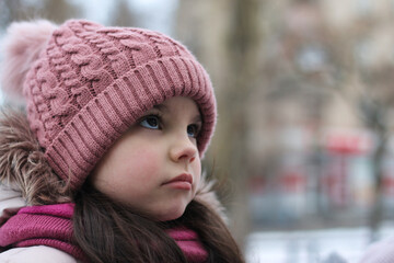 Little smiling white girl with long dark hair in a knitted hat on a background of snow and blurred buildings