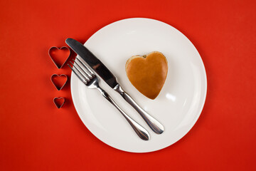 A heart-shaped burger on a white plate with a knife and fork on a red background. The view from the top. The concept of Valentine's Day.