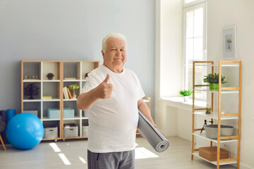 Happy senior man at home with sports mat in hand giving thumbs-up and looking at camera