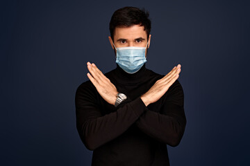 Young guy in black sweater and face mask isolated on Pacific Blue background studio portrait. Pandemic coronavirus 2019-ncov sars covid-19 flu virus concept. Showing stop gesture with crossed hands