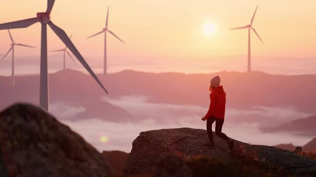 Young woman in orange jacket running up on top of mountain summit at sunset, raises arms into air, happy and drunk on life and happiness. Girl stand against landscape with wind turbines on background
